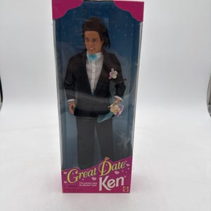 May include: A Ken doll in a clear plastic box. The doll is wearing a black tuxedo with a white shirt, a light blue bow tie, and holding a small bouquet. The box has the words "Great Date Ken" printed on it.