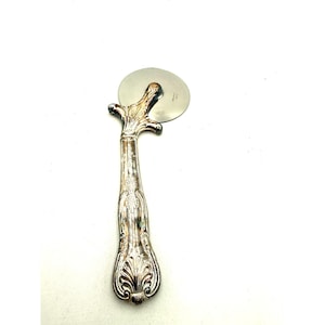 May include: A silver-coloured pizza cutter with an ornate handle. The handle has a detailed floral design and a rounded base. The circular blade is made of stainless steel. This kitchen utensil is designed for cutting pizza.