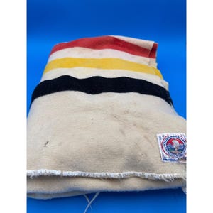 May include: A folded cream-colored blanket with horizontal stripes of red, yellow, black, and cream. A label with text and a mountain scene is visible on the lower right corner. The blanket is made of a thick, woven material.