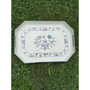 Könnte beinhalten: Ein achteckiges Serviertablett mit cremefarbener Basis und einem dunkelblauen Blumenmuster. Das Tablett zeigt ein zentrales botanisches Motiv, das von einer dekorativen Bordüre umgeben ist, und verfügt über zwei integrierte Seitengriffe.