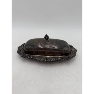 May include: An oval silver-toned butter dish with a removable lid featuring a central finial handle. The base has an ornate, raised decorative border with leaf-like patterns, and the metal surface shows signs of natural tarnish and wear.