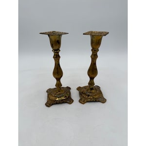 Puede incluir: Dos candeleros antiguos de latón. Cada candelero tiene una base cuadrada con detalles ornamentados, un tallo delgado y curvo y una parte superior decorativa. El latón tiene una pátina cálida y envejecida, lo que sugiere que son piezas antiguas. Los candeleros están sobre un fondo blanco.
