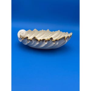 May include: A white, seashell-shaped decorative bowl with a gold-coloured trim. The bowl has a textured surface and a wavy edge. The bowl is set against a solid blue background. This decorative bowl is ideal for coastal decor.