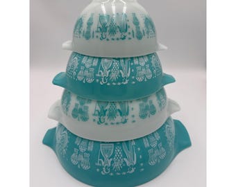 May include: A set of four stacked glass mixing bowls with handles. The bowls alternate between solid white and solid teal colors. Each bowl features a repeating white graphic print of figures, pineapples, and various kitchen utensils.