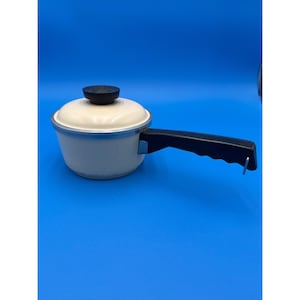 May include: A small, off-white saucepan with a matching lid and a black handle. The lid has a black knob. The pan has a silver rim and a black handle. The saucepan is designed for cooking and is suitable for kitchen use.