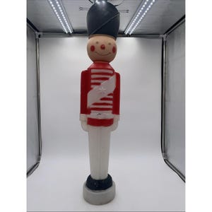 Puede incluir: Decoración vintage de soldado de juguete. El soldado viste una chaqueta roja con rayas blancas y una faja blanca. Tiene un sombrero negro y una cara sonriente. La base es gris y negra.