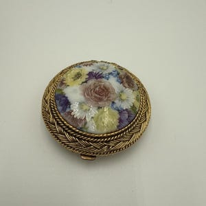 以下が含まれることがあります： ゴールド調の編み込み模様の縁取りが施された円形の金属製コンパクトケース。中央には、ピンク、黄色、紫の色合いのバラ、デイジー、その他の小さな花々が描かれた円形の磁器製インレイがはめ込まれています。