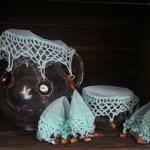 Op de afbeelding: Een set gehaakte deksels voor glazen potten en een kan. De deksels zijn gemaakt van lichtblauwe gehaakte kant en hebben een delicate, vrouwelijke uitstraling. De deksels zijn perfect om voedsel of dranken te beschermen tegen insecten en stof.