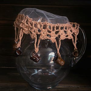 May include: A glass pitcher with a crocheted cover and brown ceramic charms hanging from the bottom. The cover is made of a fine mesh material and has a crocheted border.