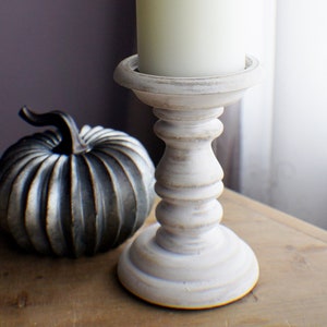 Könnte beinhalten: Weiße Kerze auf einem weißen Holzkerzenhalter mit einem silbernen Kürbis im Vordergrund.