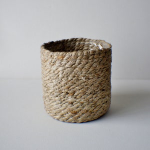 Può includere: Un cesto cilindrico realizzato con fibre di iuta naturale intrecciate. Il cesto presenta una superficie strutturata e una palette di colori neutri, adatta a vari stili di arredamento. La parte superiore aperta consente un facile accesso e la visualizzazione del contenuto.