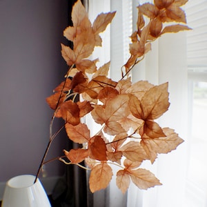 May include: A white ceramic vase with a brown leafy branch with dried leaves. The vase is sitting on a wooden table.