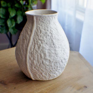 May include: A bulbous ceramic vase with a speckled off-white finish and a heavily textured, crinkled surface. The vase features a distinct vertical raised seam running down the side and a wide, open circular mouth at the top.
