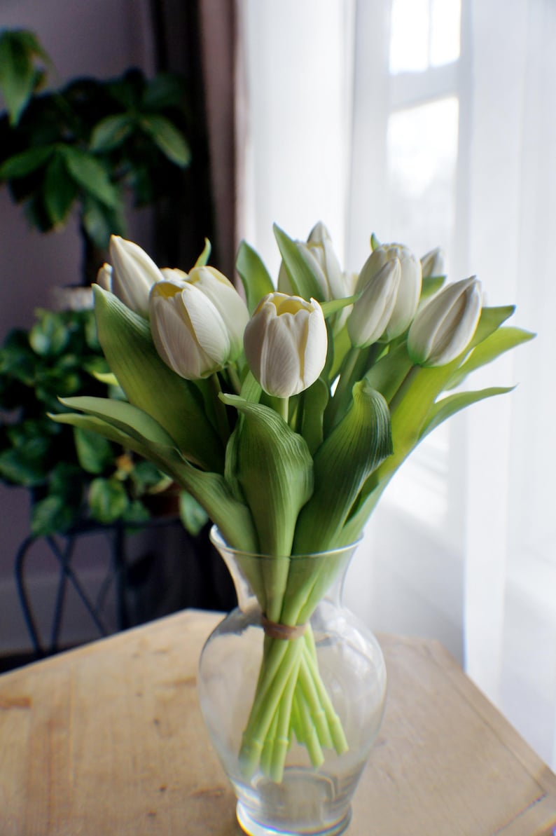 May include: A bunch of white tulips in a clear glass vase. The flowers have creamy white petals and green leaves. The vase is tied with twine and sits on a wooden table, with a window in the background.