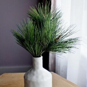 May include: A white ceramic vase with the word "BLOOM" in black lettering, holding a bundle of green pine needles. The vase sits on a wooden surface, with a window and curtain in the background.