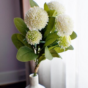 May include: A bouquet of white and green artificial flowers in a white ceramic vase. The flowers are arranged in a cluster and have a realistic appearance.
