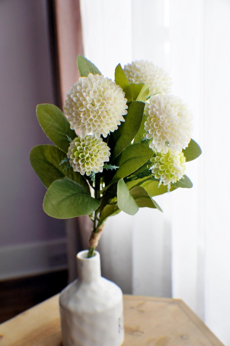May include: A bouquet of white and green artificial flowers in a white ceramic vase. The flowers are arranged in a cluster and have a realistic appearance.