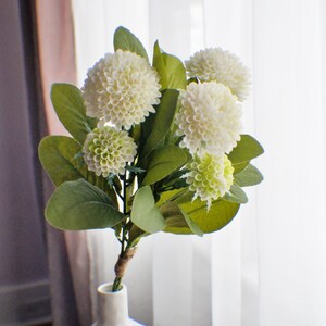 May include: A bouquet of white and green artificial hydrangea flowers in a white ceramic vase. The vase has a textured surface and is sitting on a wooden table.