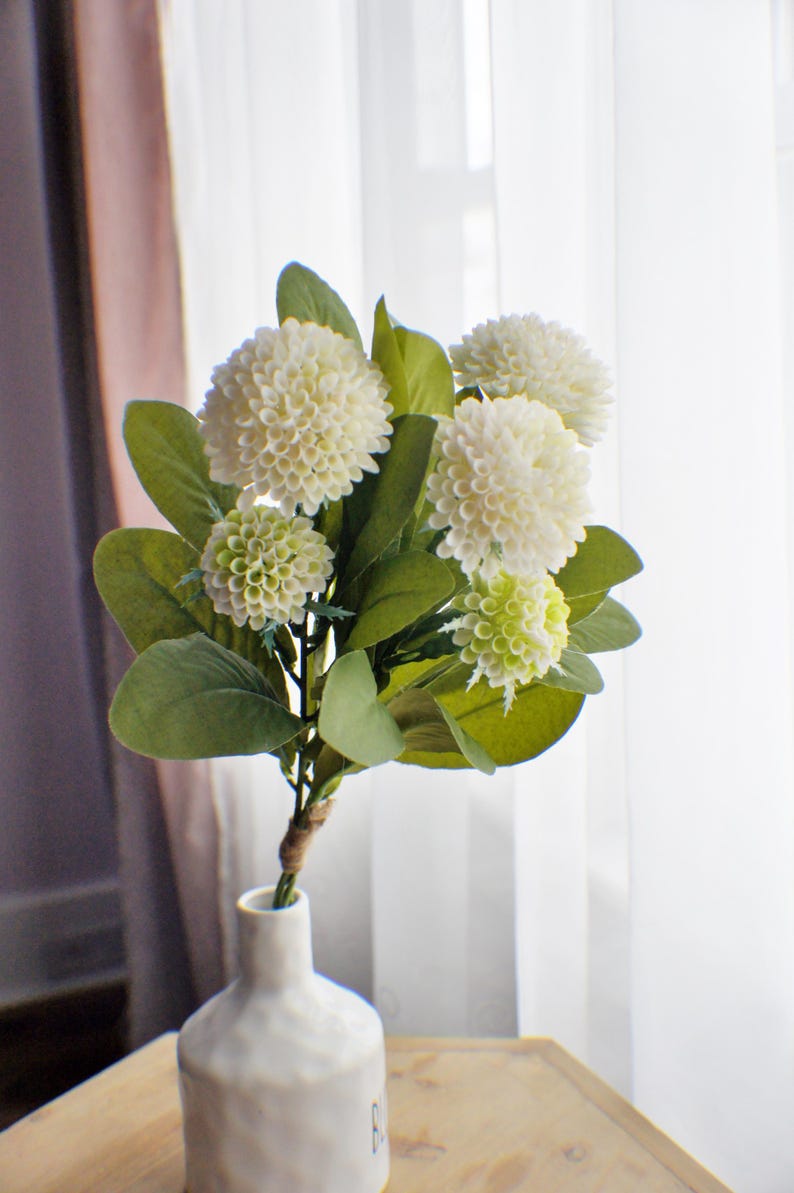 May include: A bouquet of white and green artificial hydrangea flowers in a white ceramic vase. The vase has a textured surface and is sitting on a wooden table.