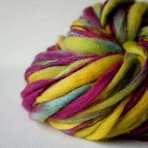 May include: A close-up of a vibrant ball of hand-dyed roving yarn. The yarn features bold colors including yellow, fuchsia, teal, and green. The wool is thick and soft, perfect for fiber art projects.