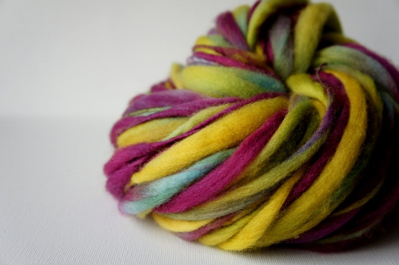 May include: A close-up of a vibrant ball of hand-dyed roving yarn. The yarn features bold colors including yellow, fuchsia, teal, and green. The wool is thick and soft, perfect for fiber art projects.