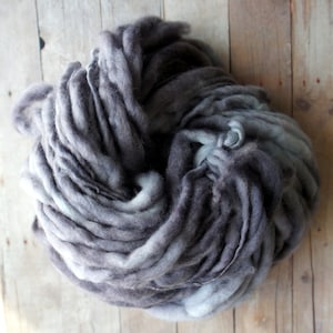 Puede incluir: Un ovillo de hilo teñido a mano en tonos grises y blancos. El hilo está hecho de fibras suaves y esponjosas y es perfecto para tejer o hacer ganchillo.