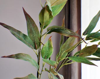 May include: A white vase holds a decorative plant with long, green leaves and reddish-brown tips. The leaves have a textured appearance, and the plant's stems are a dark brown color. The background is a neutral gray and brown.