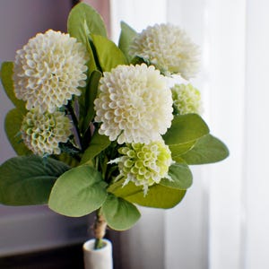 May include: A bouquet of white hydrangea flowers with green leaves in a white ceramic vase.