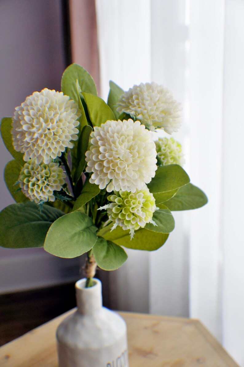 May include: A bouquet of white hydrangea flowers with green leaves in a white ceramic vase.