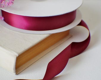 May include: A stack of white spools of burgundy ribbon, with a length of ribbon unfurling. A small, off-white book is beneath the spools. Lavender sprigs are in the upper left corner. The background is white.