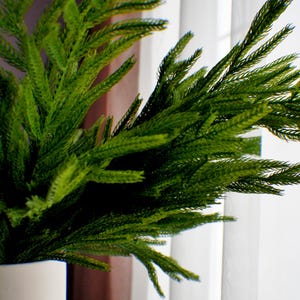 May include: A green artificial plant with long, thin leaves in a white vase. The plant is placed in front of a white sheer curtain.