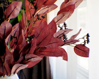 Könnte beinhalten: Eine weiße Vase enthält ein Arrangement aus künstlichem Eukalyptus. Die Blätter sind tief burgunderfarben mit detaillierten Adern, ergänzt durch kleine, dunkle Beeren. Das Arrangement steht vor einem hellen Fenster.