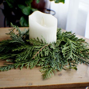 May include: A white LED candle sits atop a circular evergreen wreath. The candle has a realistic flame effect. The wreath is made of fresh, green cedar branches. The arrangement is on a wooden surface, creating a natural, rustic aesthetic.