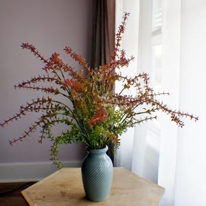 May include: A bouquet of artificial flowers in a light blue vase sits on a wooden hexagonal table. The flowers have green, orange, and red hues. The vase has a textured surface. A window with white curtains is in the background.