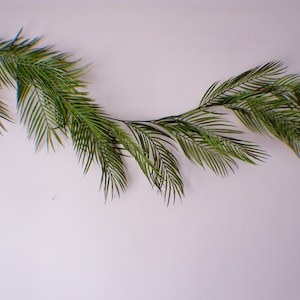 May include: A long, artificial palm leaf garland with multiple green fronds. The garland is arranged in a curved shape against a white background, creating a natural, tropical aesthetic. Ideal for home decor or event styling.