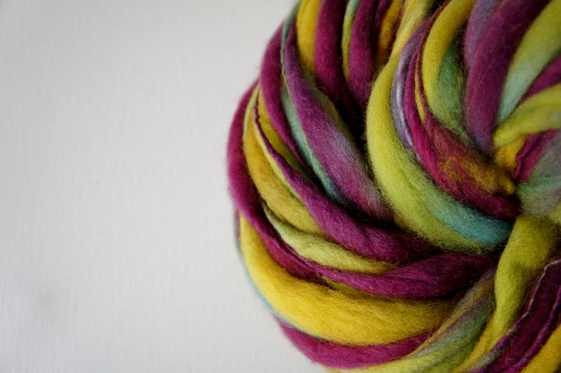 May include: A close-up of a vibrant skein of hand-dyed yarn. The yarn features a blend of colors, including yellow, purple, green, and teal. The wool is thick and textured, ideal for knitting or other fiber crafts.