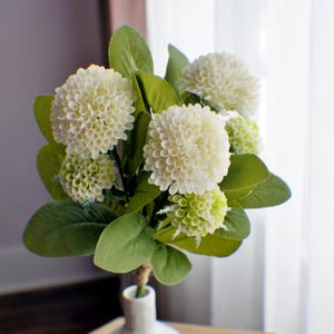 May include: A bouquet of white and green artificial flowers in a white ceramic vase with the word "bloom" written on the bottom. The vase is sitting on a wooden table.