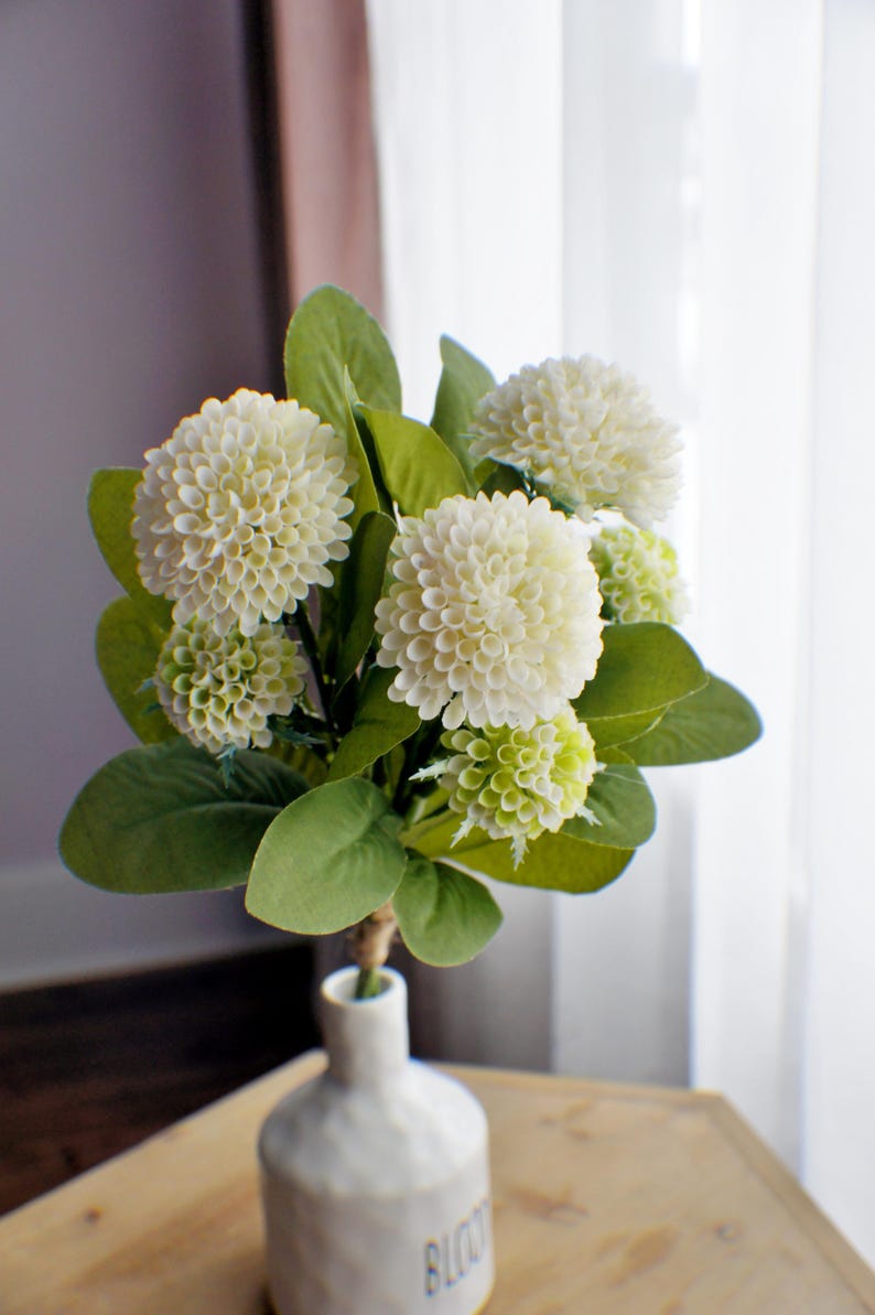 May include: A bouquet of white and green artificial flowers in a white ceramic vase with the word "bloom" written on the bottom. The vase is sitting on a wooden table.