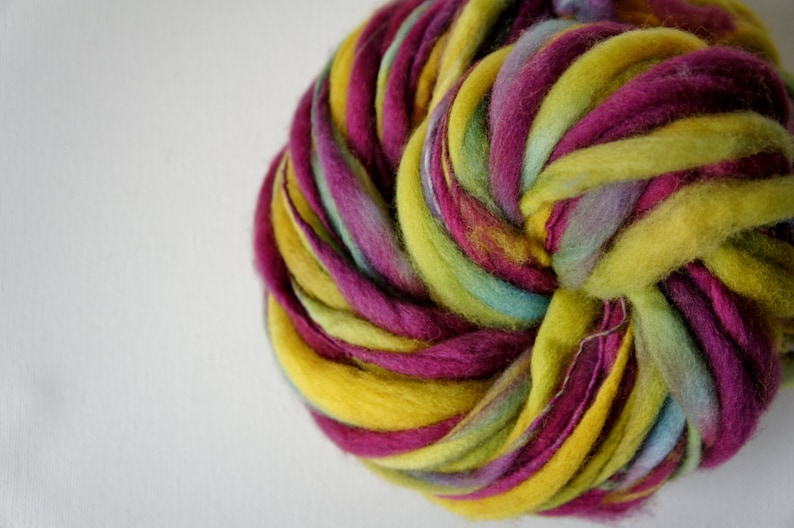 May include: A close-up of a vibrant ball of hand-dyed yarn. The yarn is a mix of colors, including yellow, magenta, light green, and blue. The yarn is thick and textured, perfect for knitting or crocheting projects.