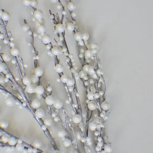 以下が含まれることがあります： 銀色の枝と白い真珠のような実が付いた、装飾用の花の茎のクローズアップ。実は枝に沿って集まっており、質感とエレガントな外観を作り出しています。休日の装飾や結婚式の装飾に最適です。