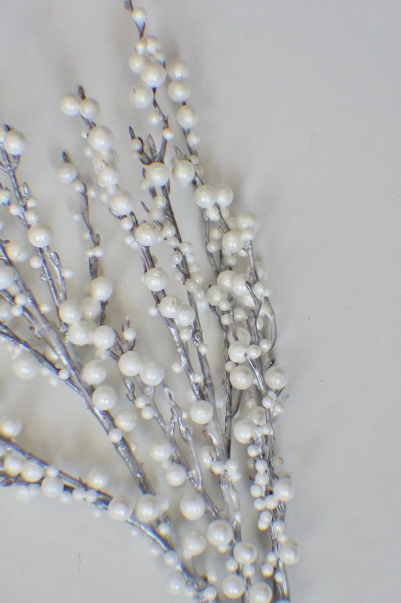 以下が含まれることがあります： 銀色の枝と白い真珠のような実が付いた、装飾用の花の茎のクローズアップ。実は枝に沿って集まっており、質感とエレガントな外観を作り出しています。休日の装飾や結婚式の装飾に最適です。
