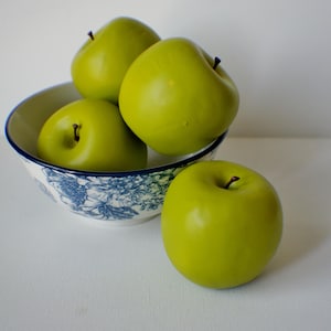 May include: A bowl with a blue and white floral pattern containing four green apples. One apple is outside the bowl. The apples are glossy with brown stems. The bowl has a blue rim.