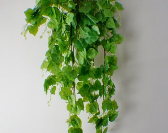 May include: A long, green artificial vine with leaves hanging down against a white wall.