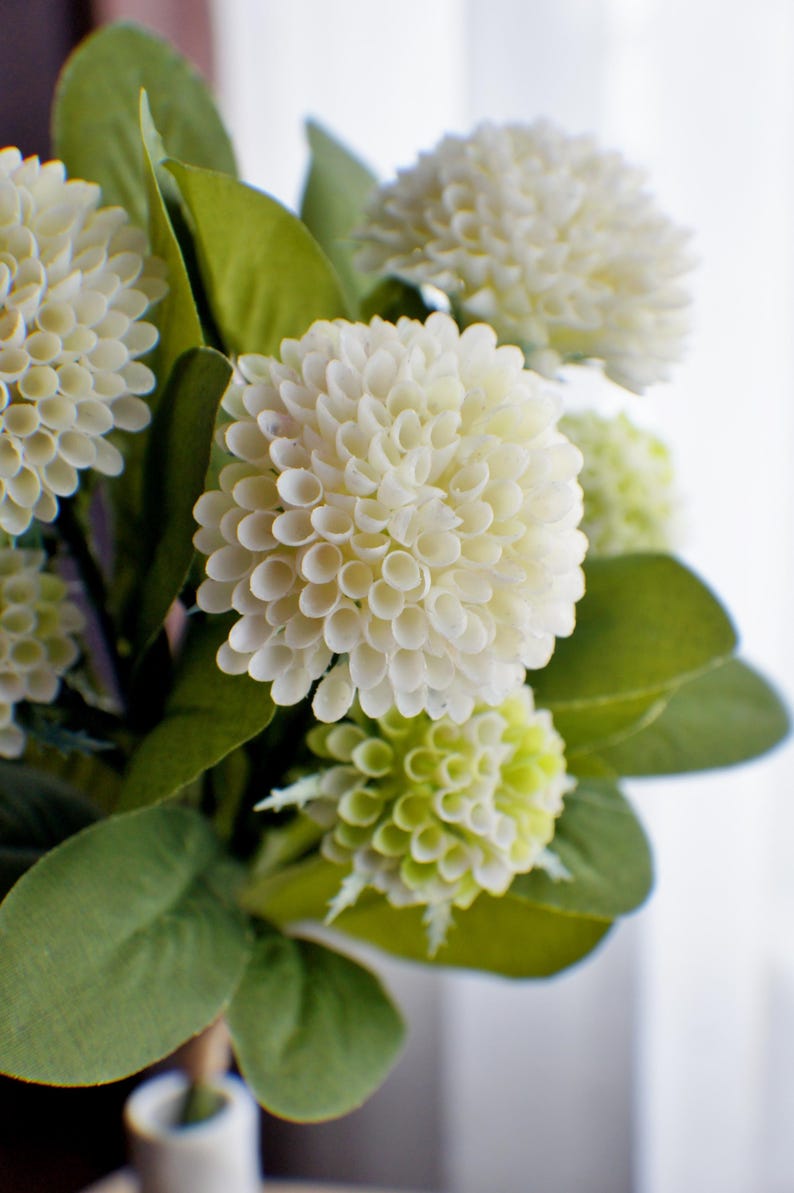 May include: A bouquet of white and green artificial flowers with green leaves. The flowers are arranged in a vase.
