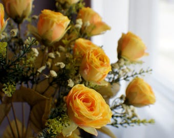 May include: A bouquet of yellow roses with green foliage. The roses are in full bloom, with layers of petals in shades of yellow and orange. The arrangement is set against a blurred background.