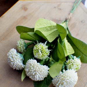 May include: A bouquet of white and green artificial flowers with green leaves. The flowers are arranged in a cluster and have a delicate, fluffy texture.
