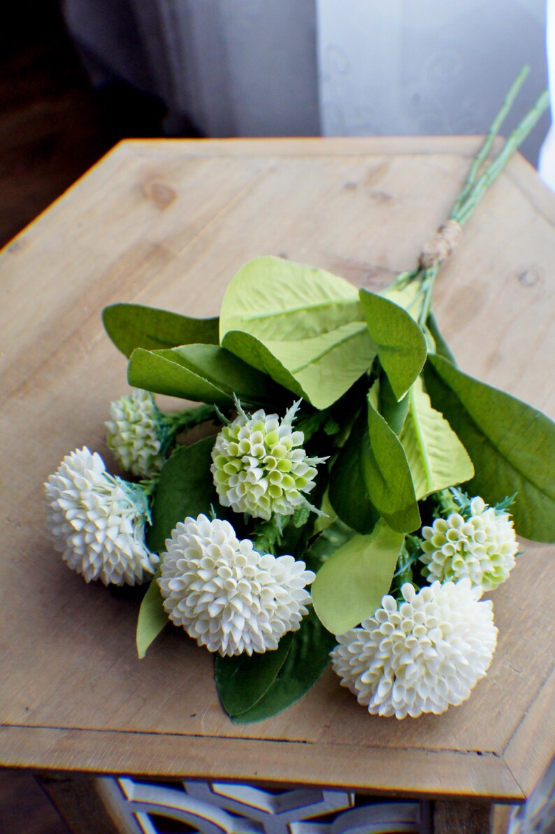 May include: A bouquet of white and green artificial flowers with green leaves. The flowers are arranged in a cluster and have a delicate, fluffy texture.