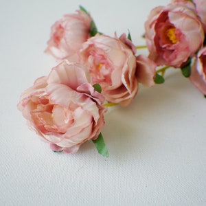 May include: A bouquet of pink silk peonies with green leaves. The peonies are arranged in a cluster, with the blooms facing outward. The background is a white surface.
