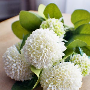 May include: A bouquet of white artificial flowers with green leaves. The flowers are arranged in a cluster and have a delicate, layered appearance.