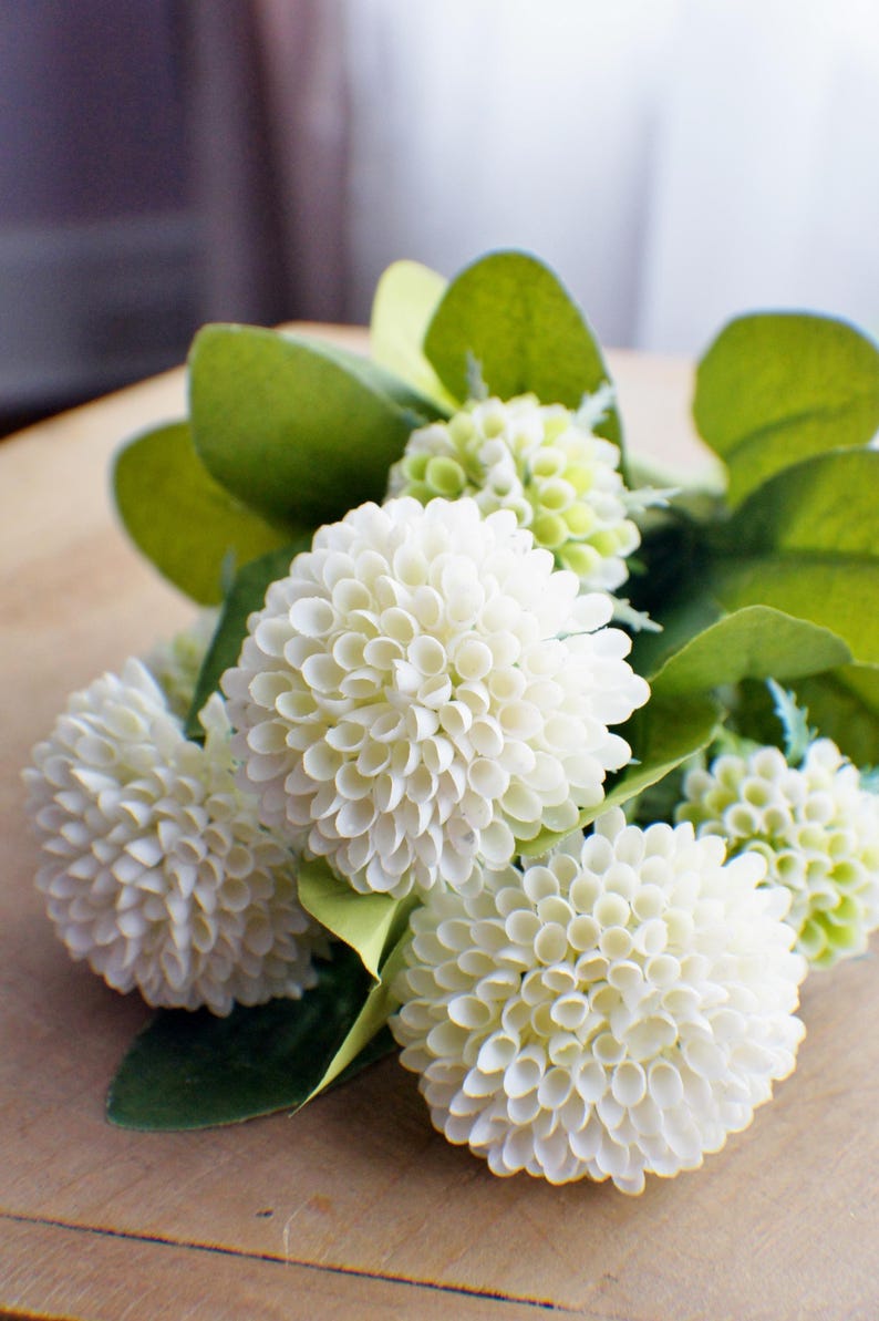 May include: A bouquet of white artificial flowers with green leaves. The flowers are arranged in a cluster and have a delicate, layered appearance.